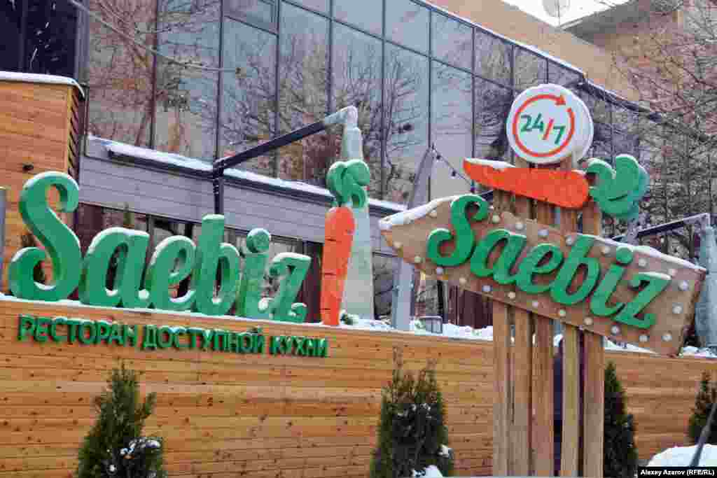 Латын қарпіндегі жазуға мемлекеттік мекемелерге қарағанда жеке меншік құрылымдар тезірек өтіп жатқандай әсер қалдырады. Латын қарпіндегі әліпбидің алғашқы нұсқасы қоғам назарына ұсынылғаннан кейін Алматыдағы мейрамханалардың бірі атауын өзгертті. Диграфты (бір дыбысты екі әріппен белгілеу) әліпби қоғамда сынға қалған болатын. Бұл нұсқаны мінегендер мысал ретінде қазақша "сәбіз" атауын&nbsp;saebiz&nbsp;деп жазу орыс тілінде дөрекі естілетін сөзді еске салатынын айтқан. Мейрамхана иесі жұрт назарын аудару үшін бұл сөзді ұтымды пайдалануды ұйғарған сыңайлы. Латын қарпінде жазылған сөздің басқа мағынасы жоқ екенін білдіру үшін сәбіздің суретін салып қойған. Атауынан өзге жазудың барлығы орысша жазылған. Жаңа әліпби нұсқаларынан диаграф қана емес, одан кейін ұсынылған апострофтар да алынып тасталғаннан кейін де бірталай уақыт дейін мейрамхана осы атауымен қалды. Суретте: Алматыдағы мейрамхана атауы. 5 желтоқсан 2018 жыл.