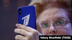 "Здоровье" көрсөтүүсүнүн алып баруучусу Елена Малышева "Биринчи каналдын" эмблемасы тагылган телефон көтөрүп турат. 